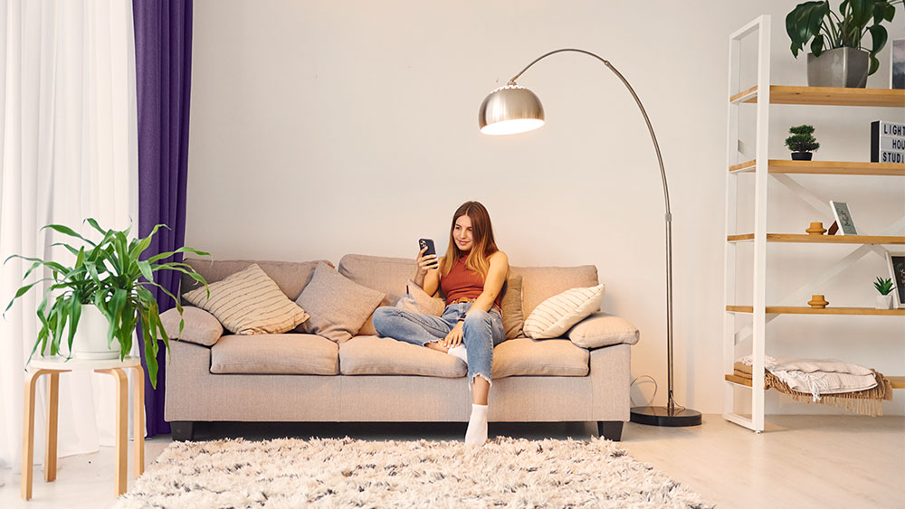 Mulher sentada no sofá de casa mexendo no celular. Luz natural entrando pela janela do lado esquerdo e um abajur ligado logo acima da mulher