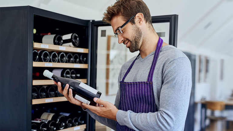 Homem analisando o rótulo de um vinho que ele pegou de uma adega para vinhos climatizada