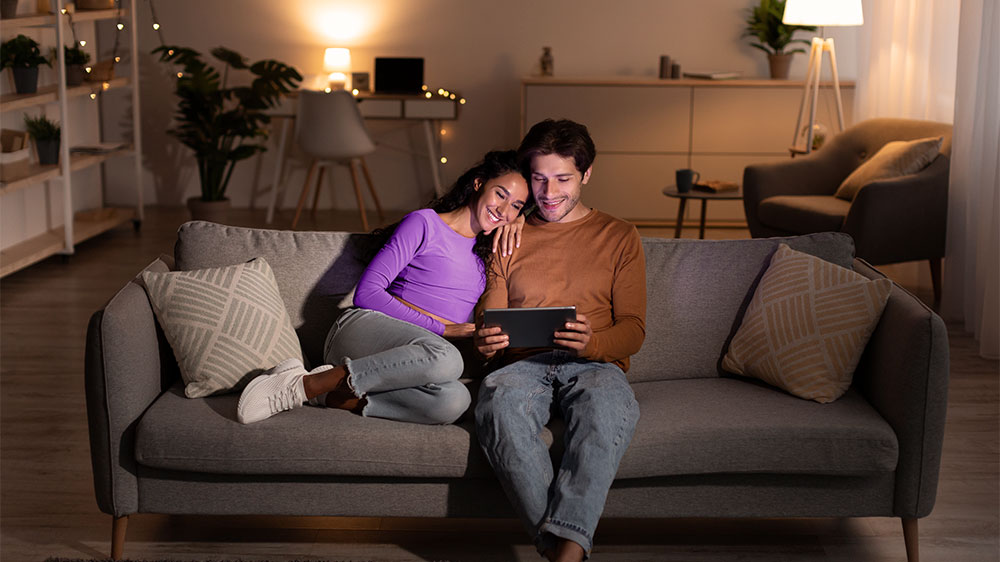 Casal relaxa em sala com iluminação eficiente, aproveitando o conforto e a economia de energia proporcionados por luminárias de LED e luzes indiretas.