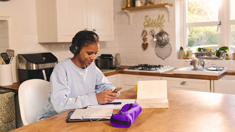 Jovem estudando com luz natural na cozinha, usando fones de ouvido e materiais escolares, economizando energia ao aproveitar iluminação do dia.