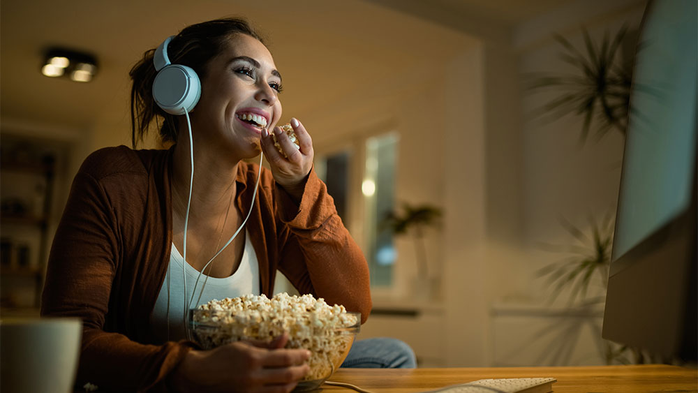 Mulher sorridente usando fones de ouvido e comendo pipoca enquanto assiste conteúdo digital em casa, demonstrando eficiência energética no entretenimento digital através de streaming em dispositivos de baixo consumo